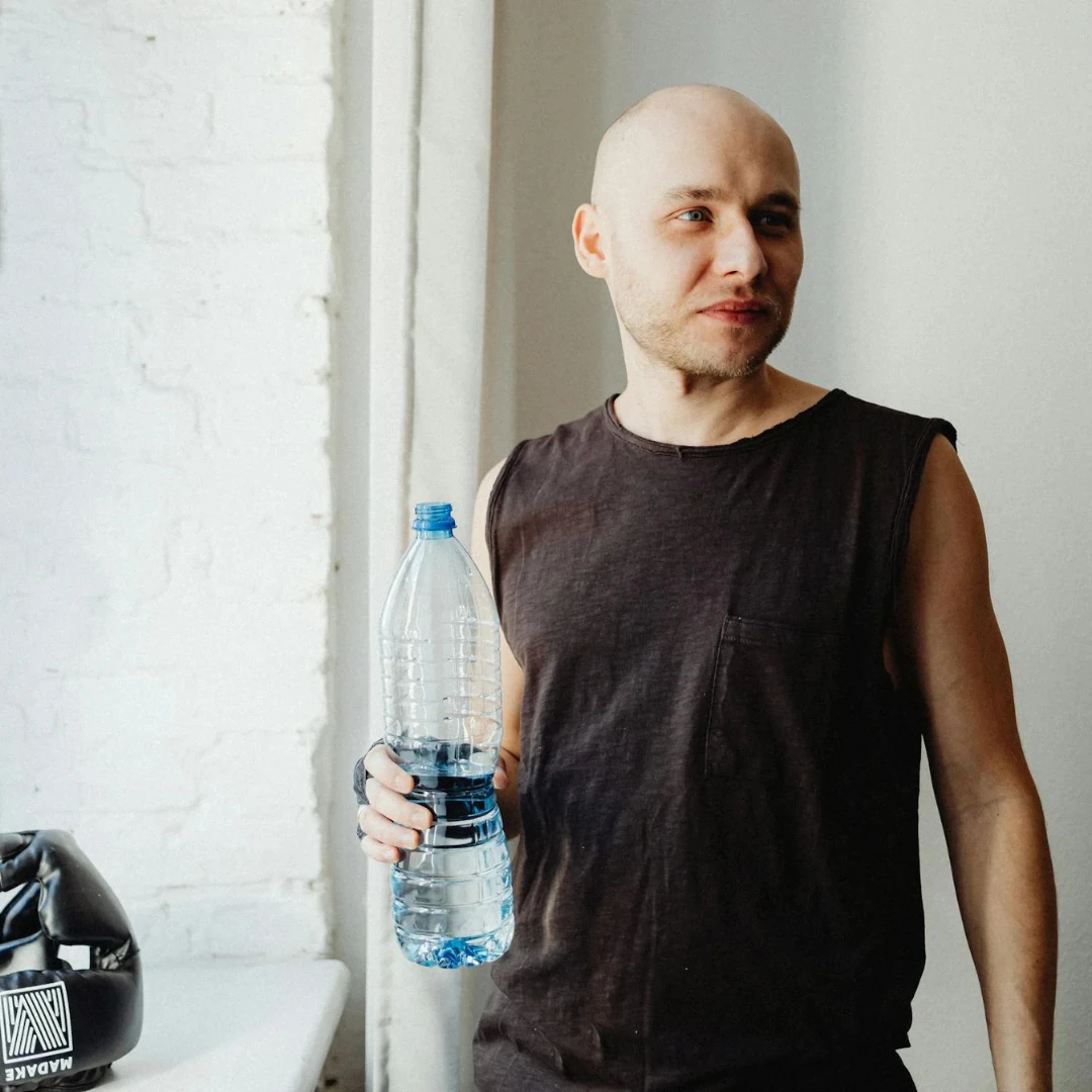 A person holding a large plastic water bottle in a well-lit indoor space, wearing a sleeveless shirt, with a relaxed expression